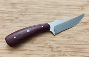 Handcrafted Purple Heart Trailing Point #285 knife with a vibrant wood handle and high-carbon steel blade on a bamboo cutting board.