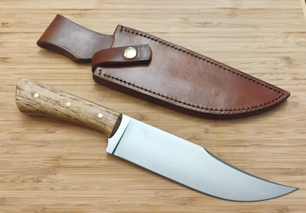 Custom Florida Pecan Upswept Clip Point, 12 5/8" Nada-Bowie, placed beside hand-cut, dyed leather sheath with retention strap on wooden